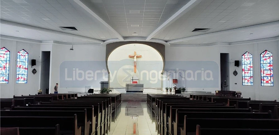 Salão principal de igreja com bancos de madeira dispostos e crucifixo acima do púlpito com vitrais ao lado