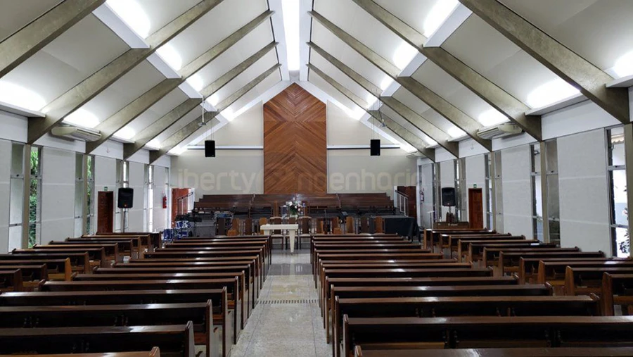 Salão Principal de Igreja bem iluminada e com equipamentos sonoros disposta com bancos de madeira