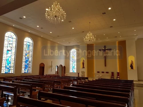 Igreja iluminada com lindos lustres e crucifixo acima do púlpito e vitrais coloridos nas paredes com bancos de madeiras dispostos no salão