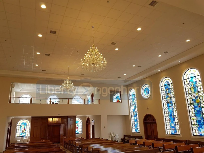 Igreja iluminada com lindos lustres e e vitrais coloridos nas paredes com bancos de madeiras dispostos no salão