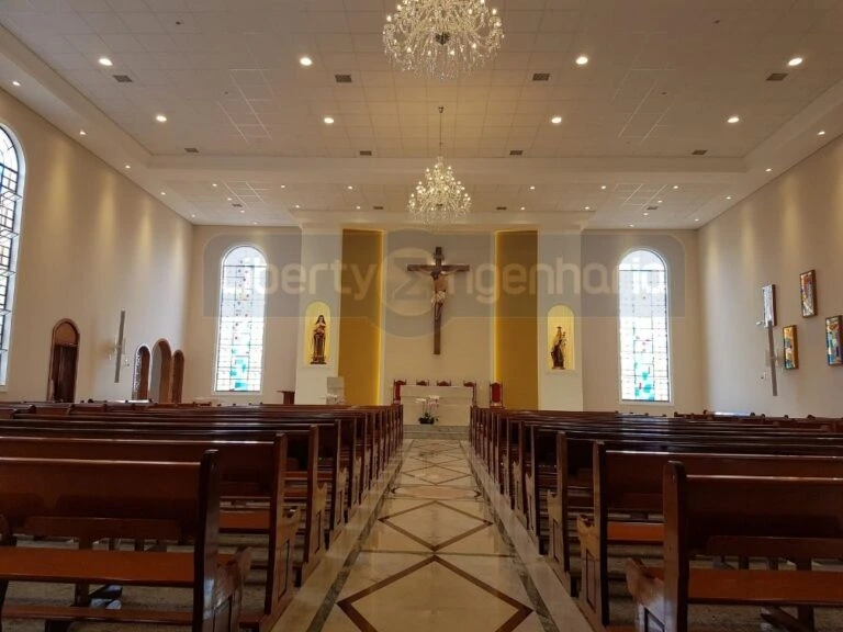 Igreja iluminada com lindos lustres e cruz cristã acima do púlpito e vitrais coloridos nas paredes com bancos de madeiras dispostos no salão