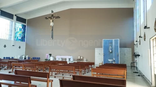 Salão de Igreja com crucifico pendurado ao teto, vitrais coloridos e bancos de madeira