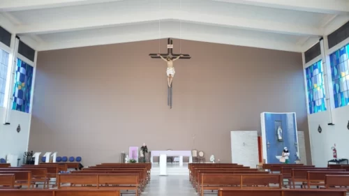 Salão de Igreja com crucifico pendurado ao teto, vitrais coloridos e bancos de madeira