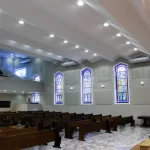 Salão principal de igreja bem iluminada com vitrais coloridos, paredes brancas e bancos de madeira